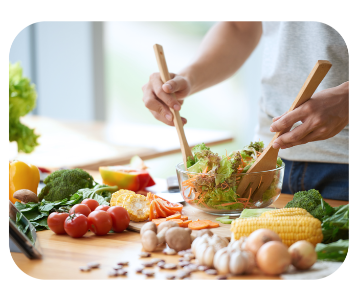 Hände halten Salatbesteck und durchmischen einen bunten Salat in einer Schüssel. Auf der Arbeitsfläche liegen verschiedene Gemüsesorten. 
