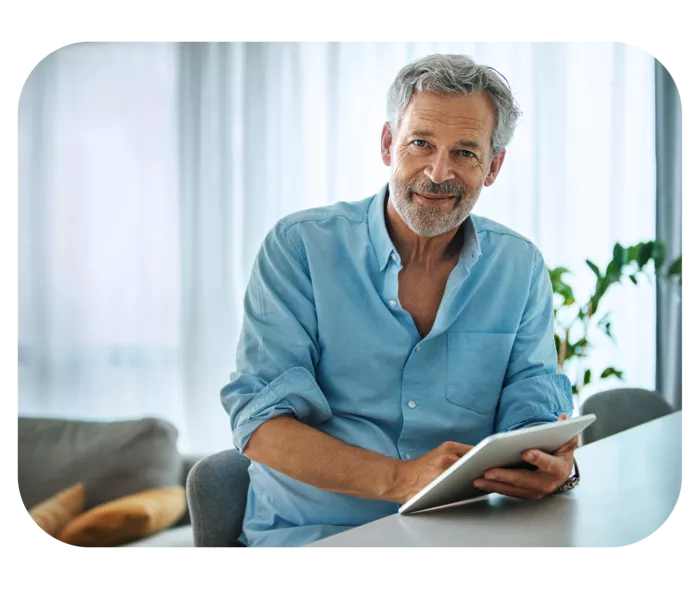 Mann mittleren Alters im blauen Hemd sitzt auf einem Stuhl, hält ein Notizbuch in der Hand und lächelt in die Kamera. 