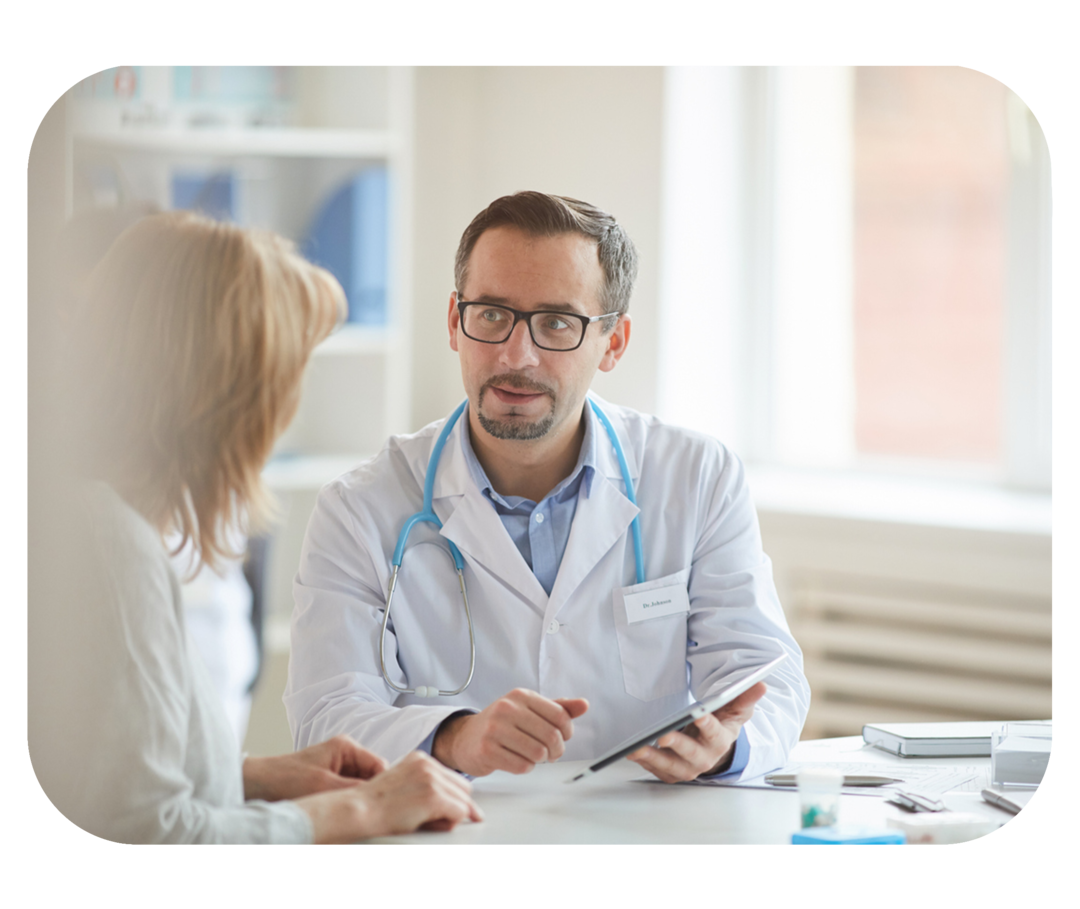 Mann mit weißem Kittel sitzt am Schreibtisch und schaut eine ihm gegenübersitzende Frau an. 