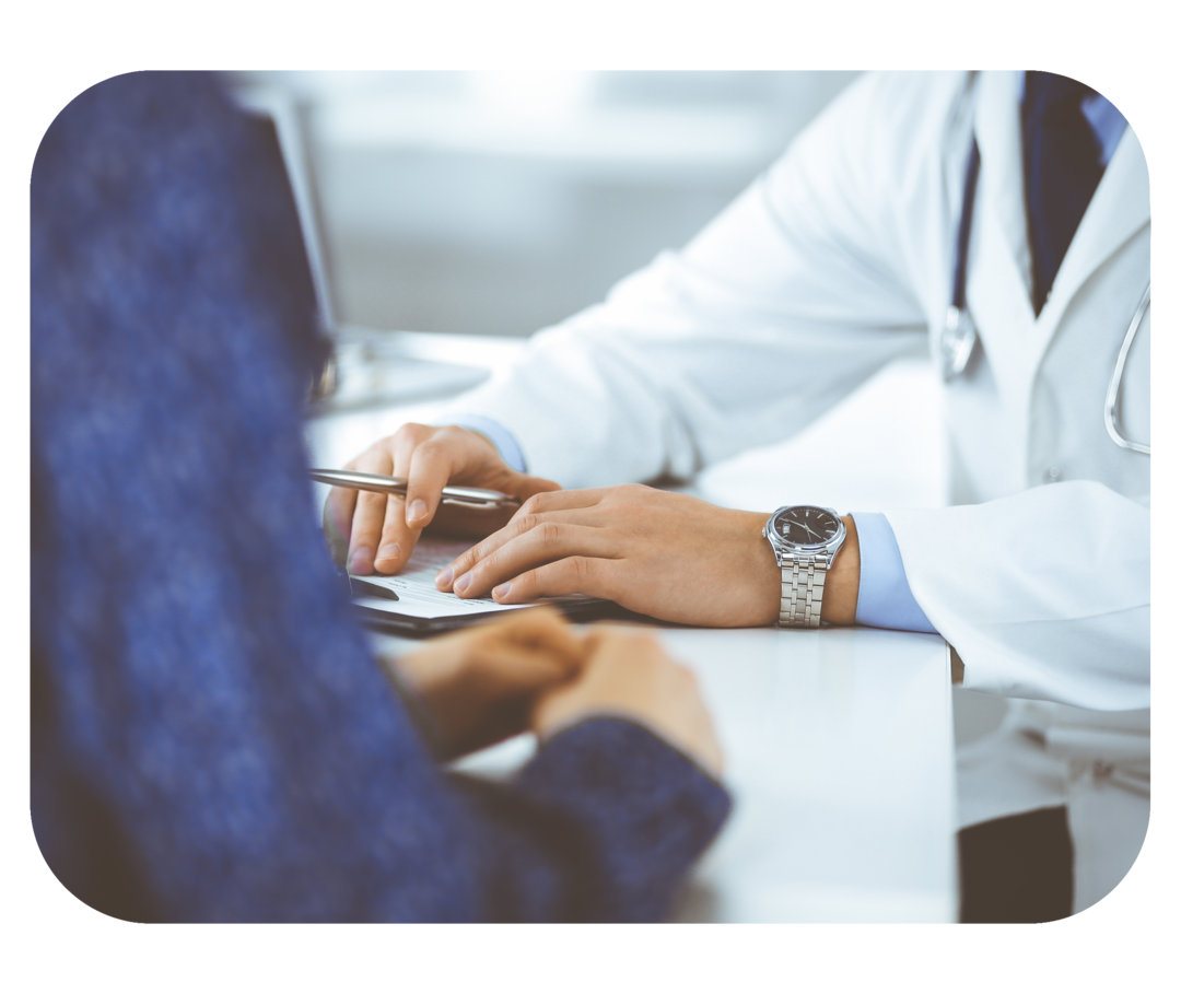 Person mit weißem Kittel sitzt an einem Tisch. Ihr gegenüber sitzt eine Person, deren Hände gefaltet auf dem Tisch liegen. 