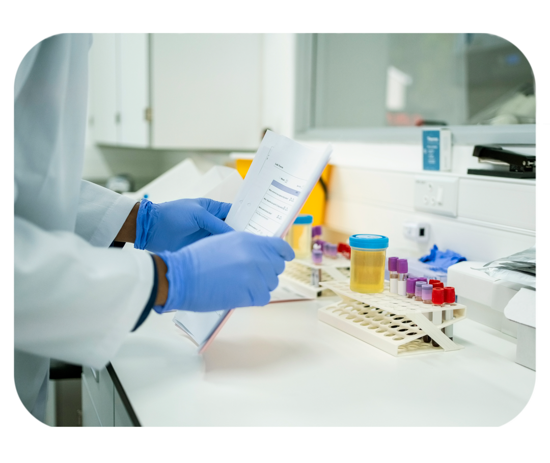 Hände in blauen Einweghandschuhen halten Zettel. Im Hintergrund Labormaterialien und ein durchsichtiger Becher mit gelber Flüssigkeit.