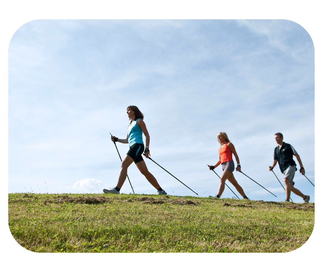 Zwei Frauen und ein Mann gehen mit großen Schritten und Nordic-Walking-Stöcken in den Händen über eine Wiese.