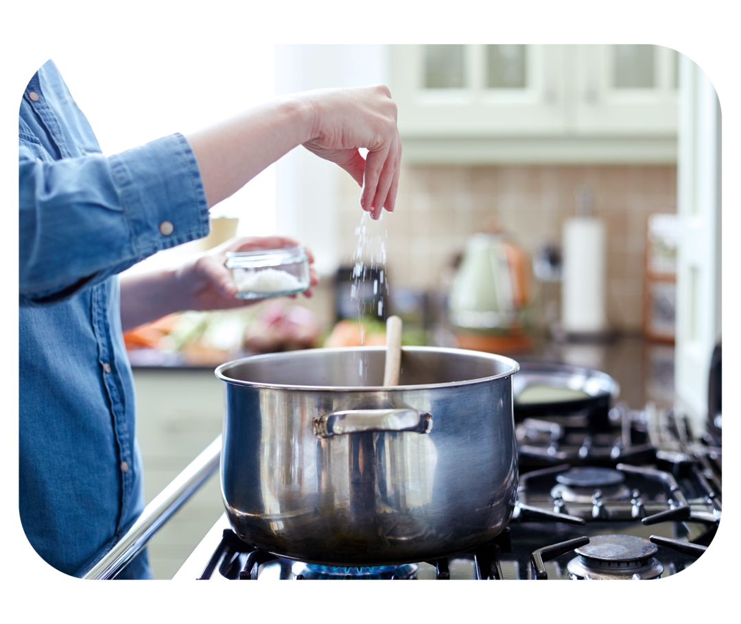 Eine Hand streut Salz in einen großen Kochtopf, der auf einem Gasherd steht. 