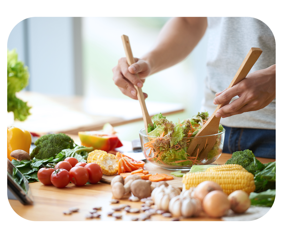 Hände halten Salatbesteck und durchmischen einen bunten Salat in einer Schüssel. Auf der Arbeitsfläche liegen verschiedene Gemüsesorten. 