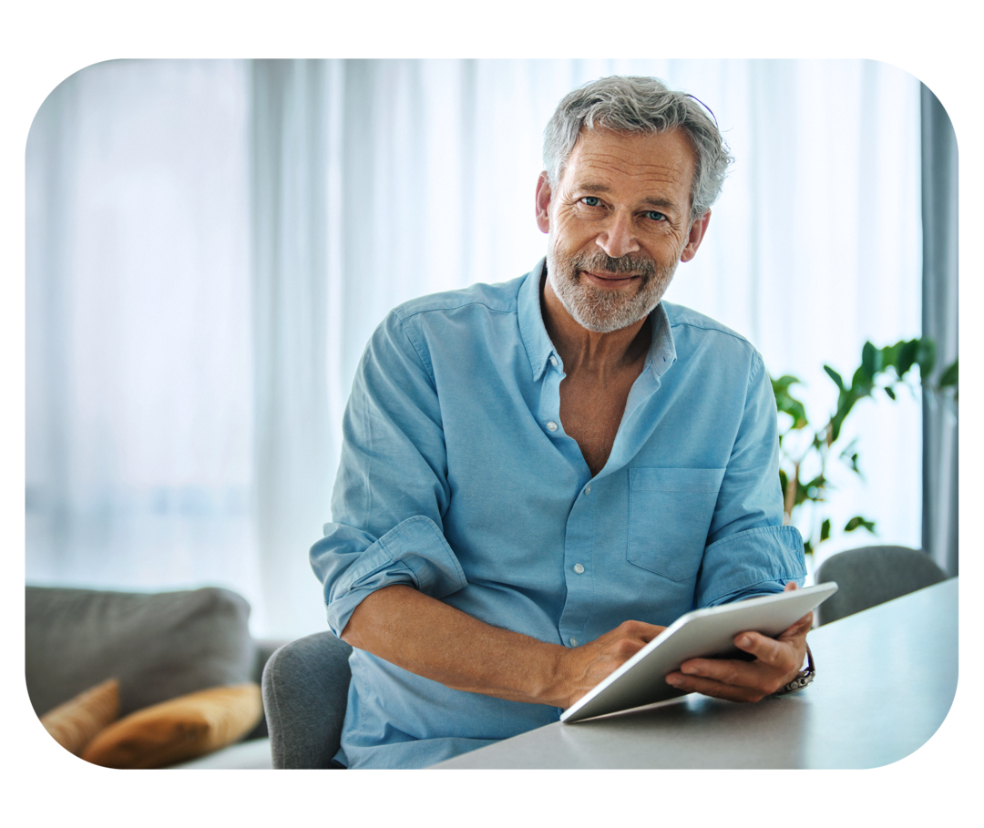 Mann mittleren Alters im blauen Hemd sitzt auf einem Stuhl, hält ein Notizbuch in der Hand und lächelt in die Kamera. 
