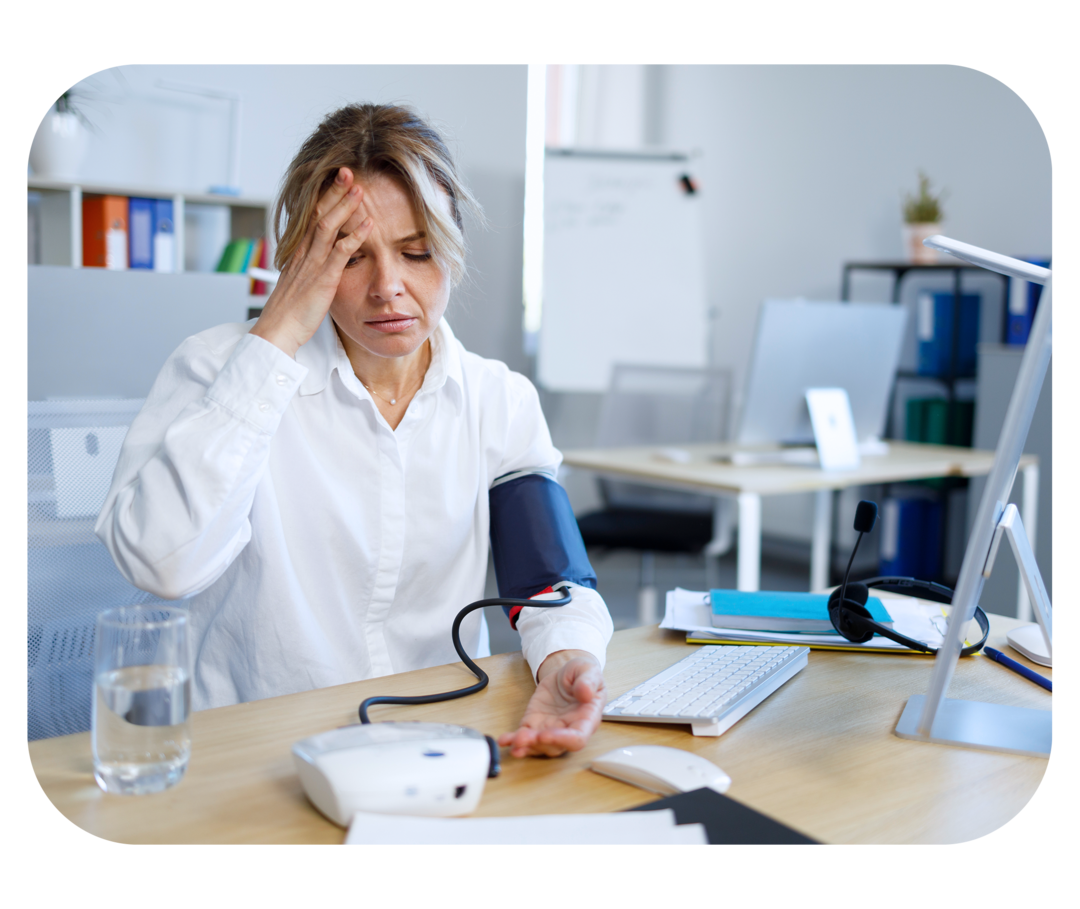 Eine Frau sitzt am Schreibtisch und greift sich mit einer Hand an den Kopf, um den anderen Arm hat sie eine Manschette eines Blutdruckmessgeräts umgebunden.