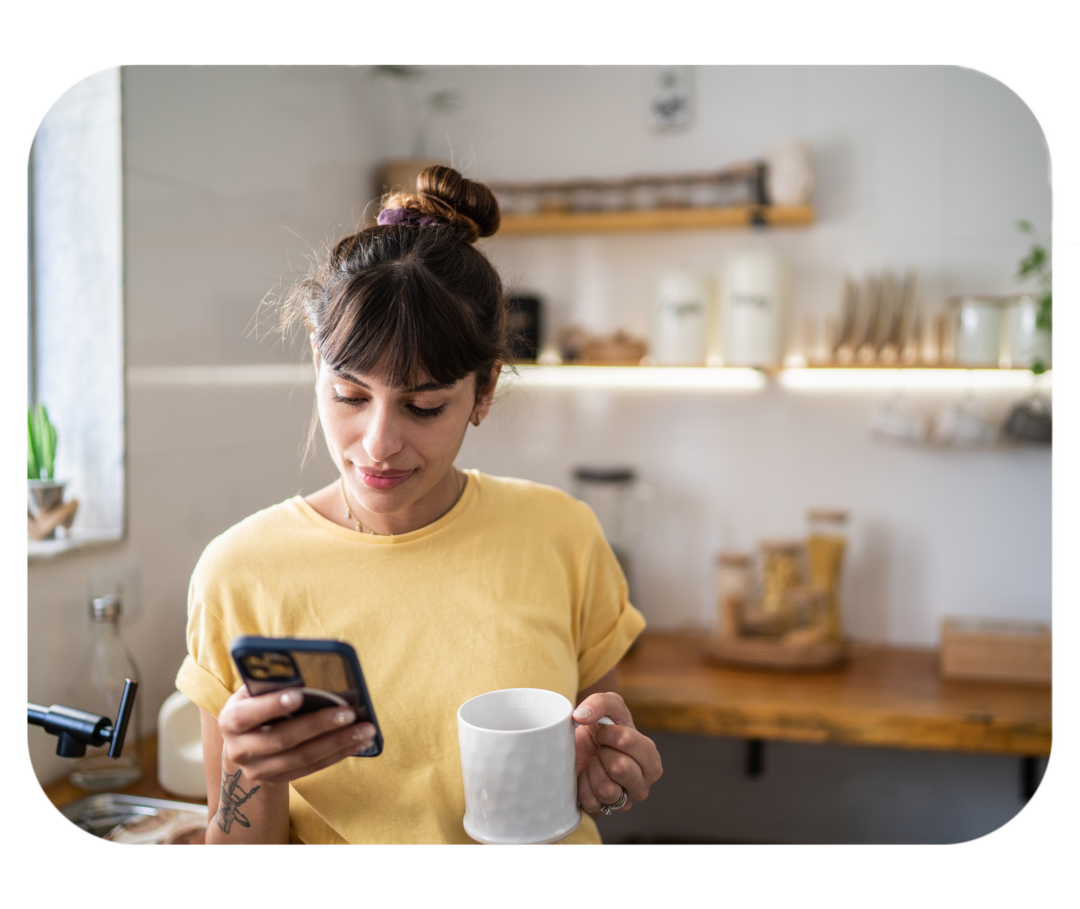 Frau mit Tasse in der Hand steht in der Küche und schaut auf ein Smartphone. 